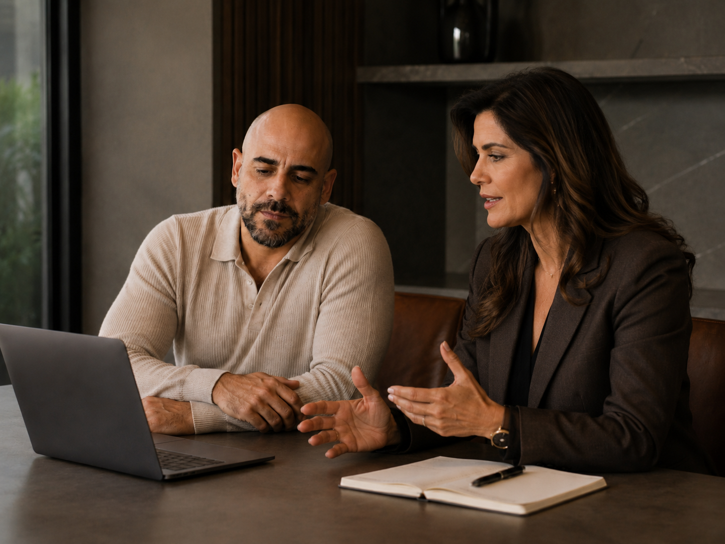 Duas pessoas conversando em uma mesa de reunião, com notebook aberto entre elas.